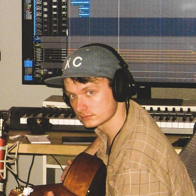 portrait of Jonathan Beard carrying a guitar in front of his computer with a keyboard on the desk.